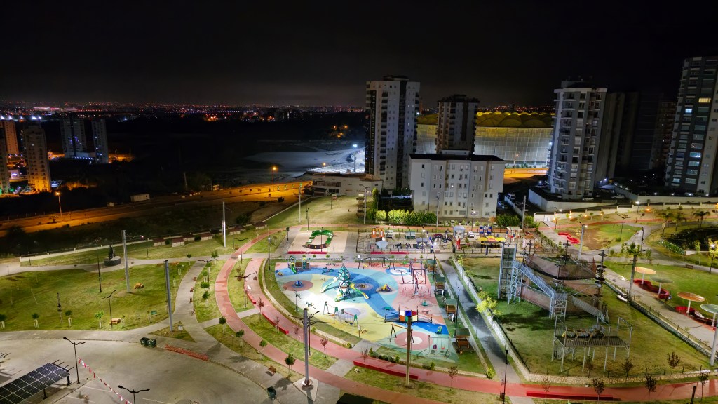 Gece manzarasında park, çocuk oyun alanı ve çevresindeki binalar.